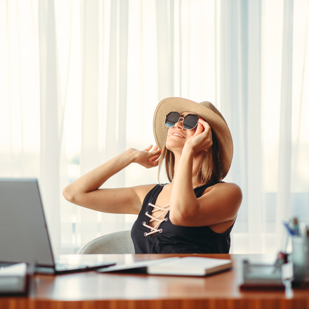 Une femme assise devant son ordinateur avec des lunettes à soleil et un chapeau de paille