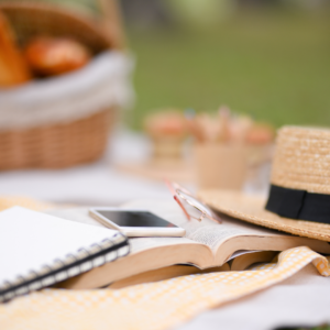 Un libre, un téléphone et un chapeau de paille sont posé dans l'herbe. Dans le fond, il y a un panier de fruit